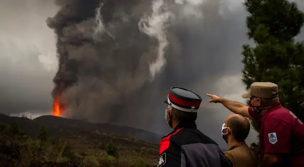 Trzej mężczyźni w maskach obserwują potężną erupcję wulkanu na La Palmie, z której wydobywa się lawa i dym. Obraz przypomina o zagrożeniu tsunami.