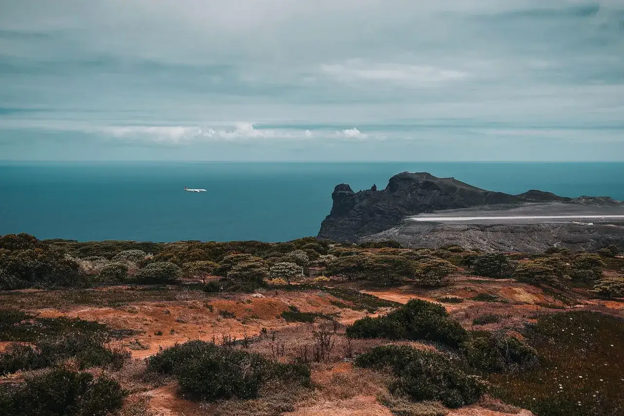 Samolot przelatuje nad oceanem w kierunku wyspy św. Heleny, gdzie widać skaliste wybrzeże i lotnisko.