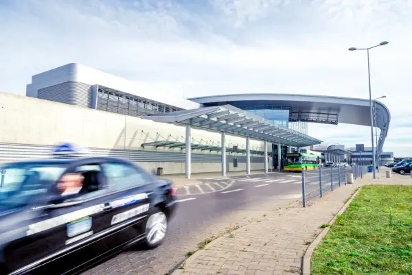 Taksówka pędzi przed nowoczesnym terminalem lotniska Poznań Ławica. W tle zielony autobus i inne pojazdy.