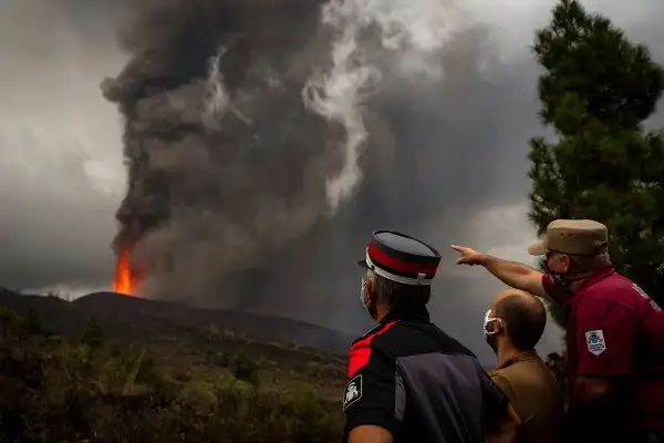 Trzej mężczyźni w maskach obserwują potężną erupcję wulkanu na La Palmie, z której wydobywa się lawa i dym. Obraz przypomina o zagrożeniu tsunami.